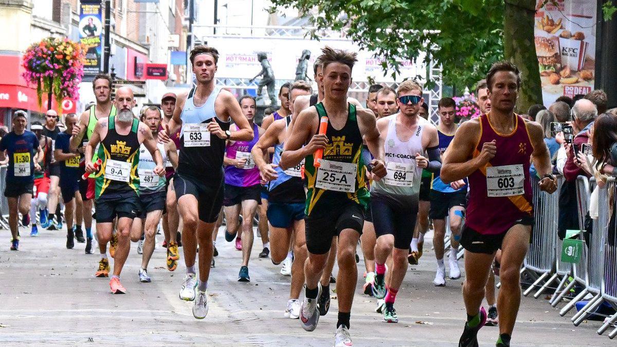 Runners heading towards the camera in the middle of a High Street. On the right is a silver barrier with spectators standing behind it.
