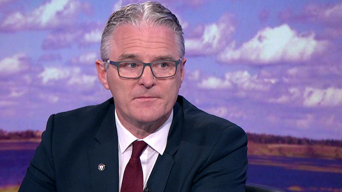 Jarlath Burns - a white/grey haired man with grey glasses on, wearing a navy suit, white shirt and maroon red tie with a GAA pin on his suit lapel