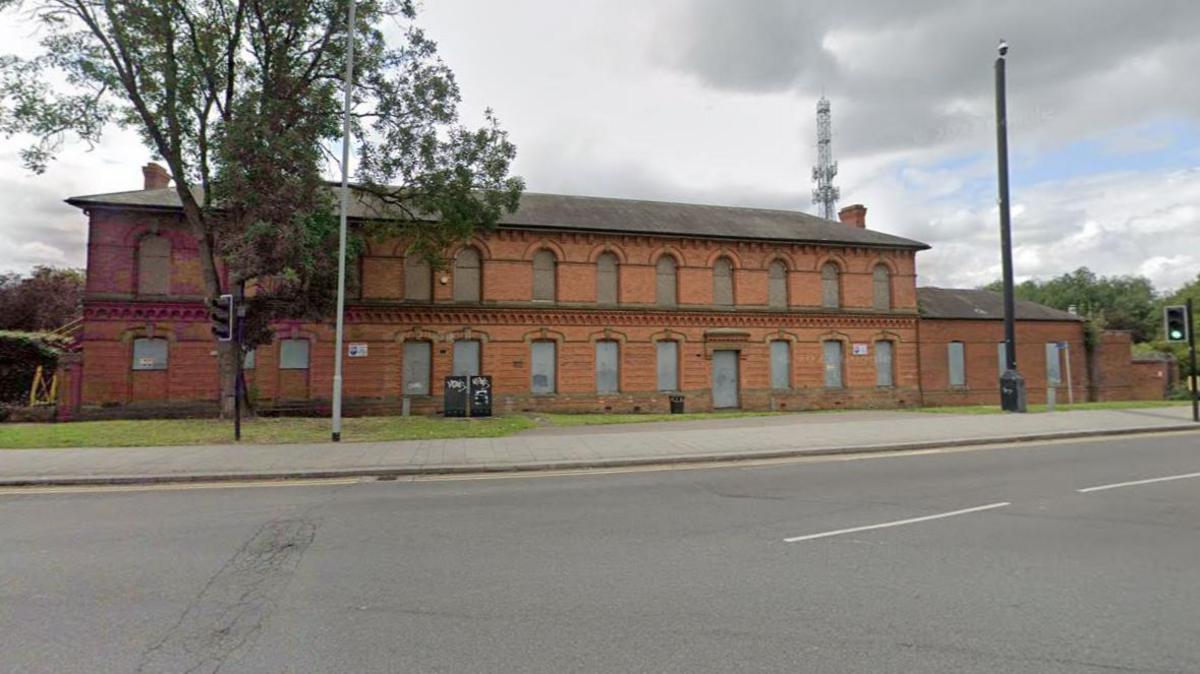 Two-storey brick building with boarded-up arched windows. There is a small grass area in front of the building and a wide road passes by.