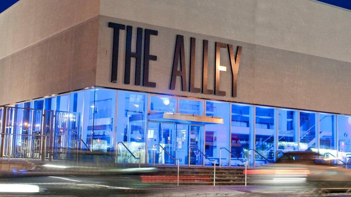 The Alley Theatre in Strabane. The theatre has its name on the front in large grey letters. The building is grey with glass panels around it on all sides. The lights inside are blue. The forefront of the picture is blurry. There are steps leading to the door of the theatre.