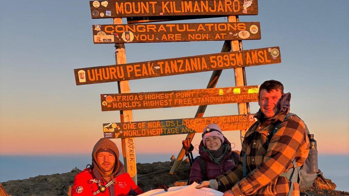 Rory, a woman and ben all stood together in front of a Mount Kilimanjaro sign. The sky is pink and yellow. The sign is brown and they are all wrapped up in thick clothing. They are all smiling at the camera and ben and rory are holding the white t-shirt.