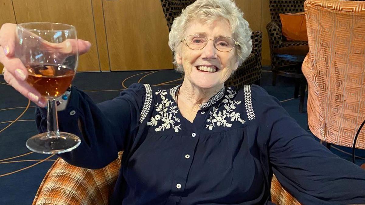 Sue pictured holding a glass of wine at the village. She has short white hair and wearing a blue top with white flowers around the collar and sat on a chair with a tartan print