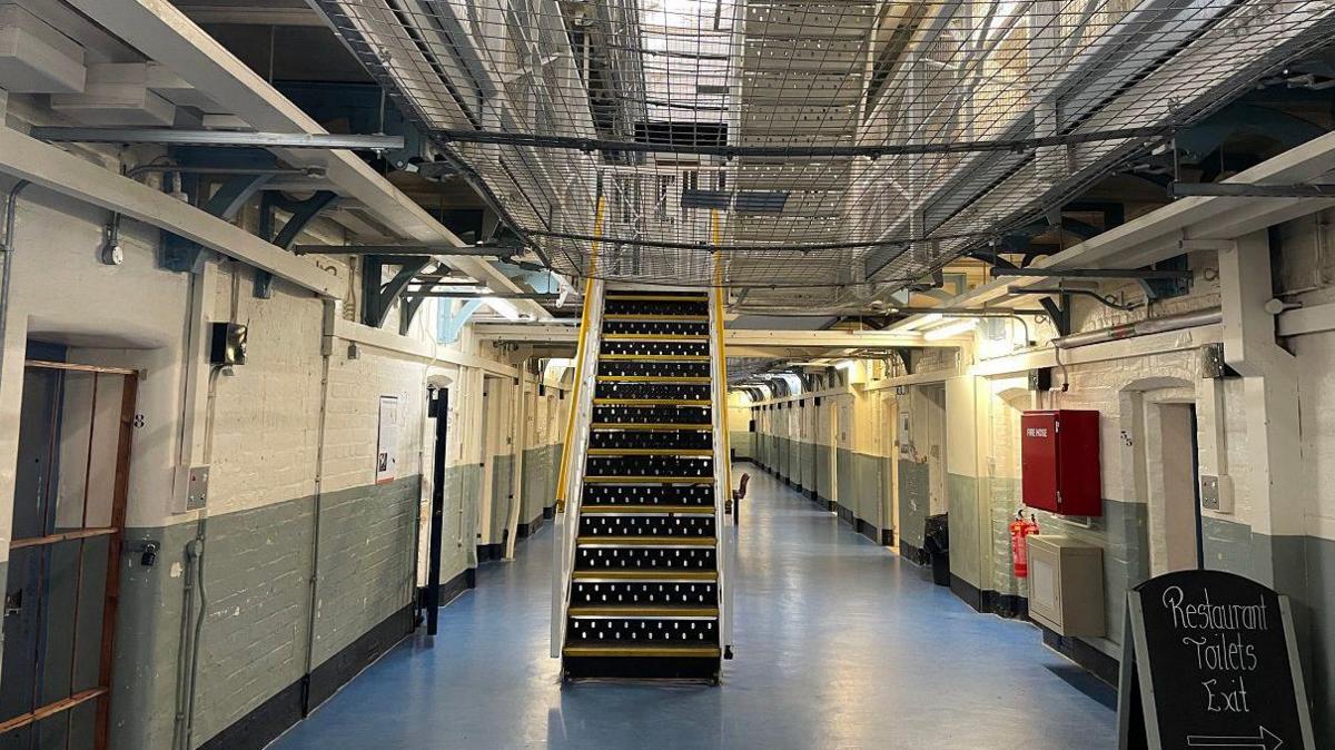 A prison corridor with doors on the left and right sides. There is a metal staircase in the centre leading to an upper floor.