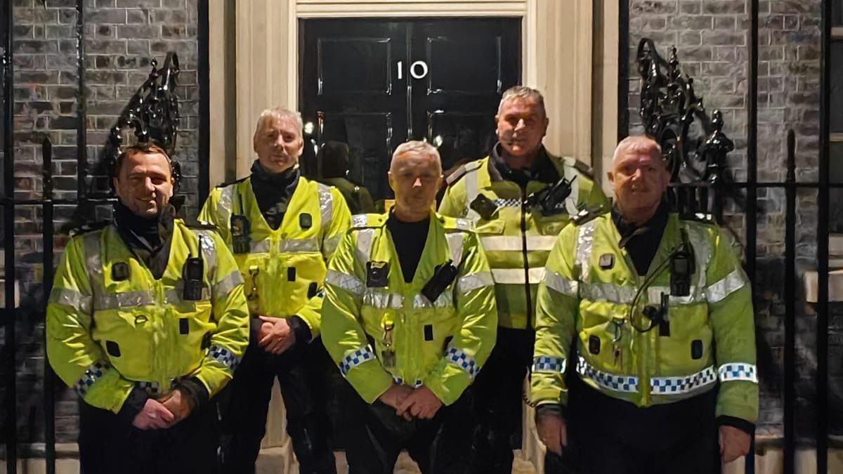 Five police officers wearing yellow are stood outside the entrance to Number 10 Downing Street, which is a black door with orante black railings either side.