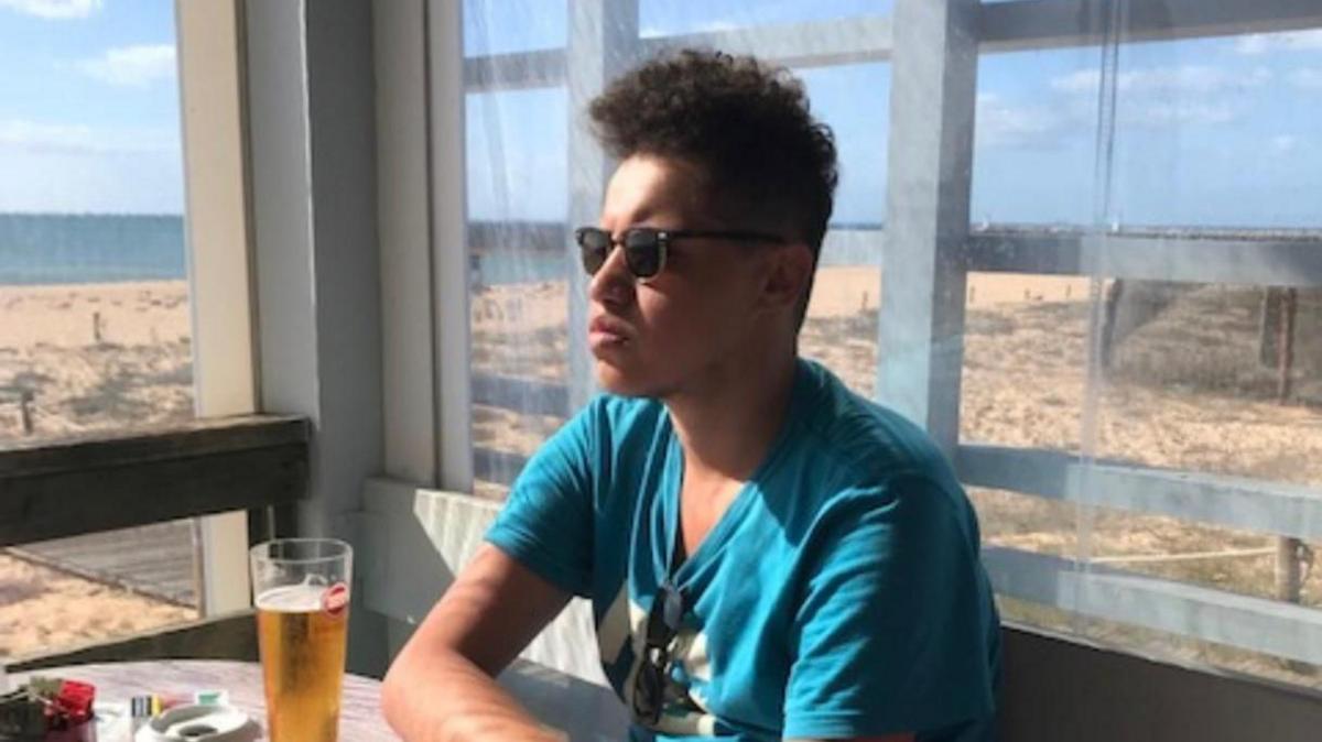 Chris Nota, who died aged 19, sits in a bar by a beach. He has a light blue t-shirt on with white writing and his sunglasses are attached to the neck of his -shirt. He has a second pair of light reflective glasses on and is looking to the left of the picture. He has a pint of beer in front of him and black curly, short hair.