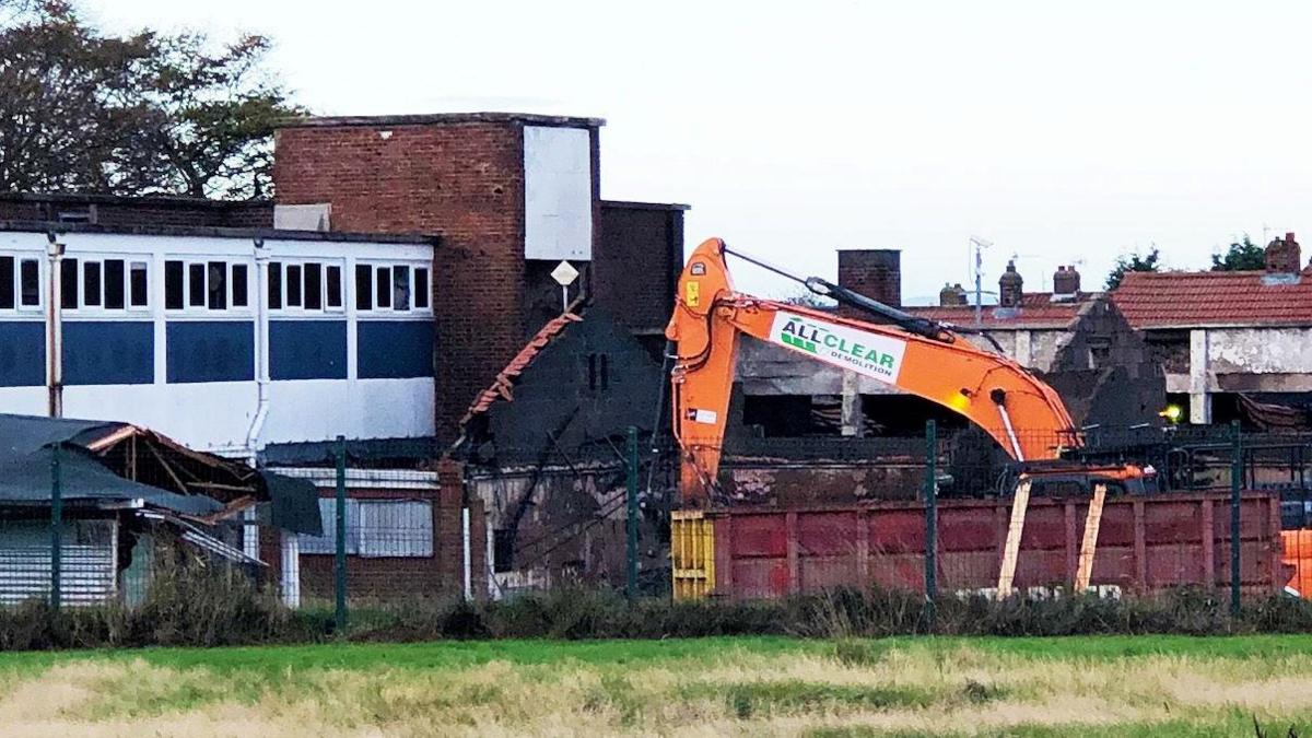 The arm and the top of the cab of an orange demolition machine can be seen behind a purple and yellow metal skip. To the left of the picture is the former school. A red brick building with a windowed section painted blue and white.