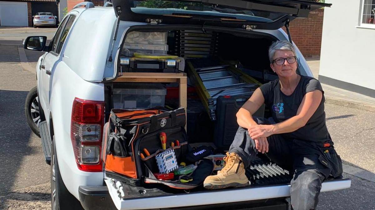 A woman sat on the open boot of her car with tools and ladders visible inside