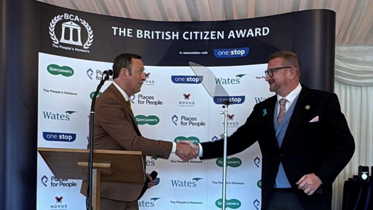 TV presenter Matt Allwright, wearing a brown suit, shakes the hand of Craig McMurrough in front of a board that reads 'The British Citizen Award'. Mr McMurrough is wearing a dark suit with blue waistcoat and white shirt. 