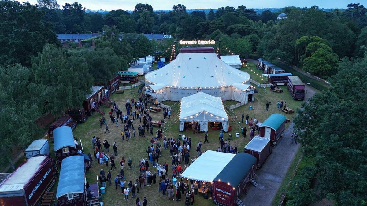 A white, domed circus tent is seen from above. It is topped with lit-up electric letters reading Gifford's Circus. The tent is pitched in a grassy field alongside a marquee and several wagons. There are several dozen people packed into the field, which is surrounded by dark green trees. 