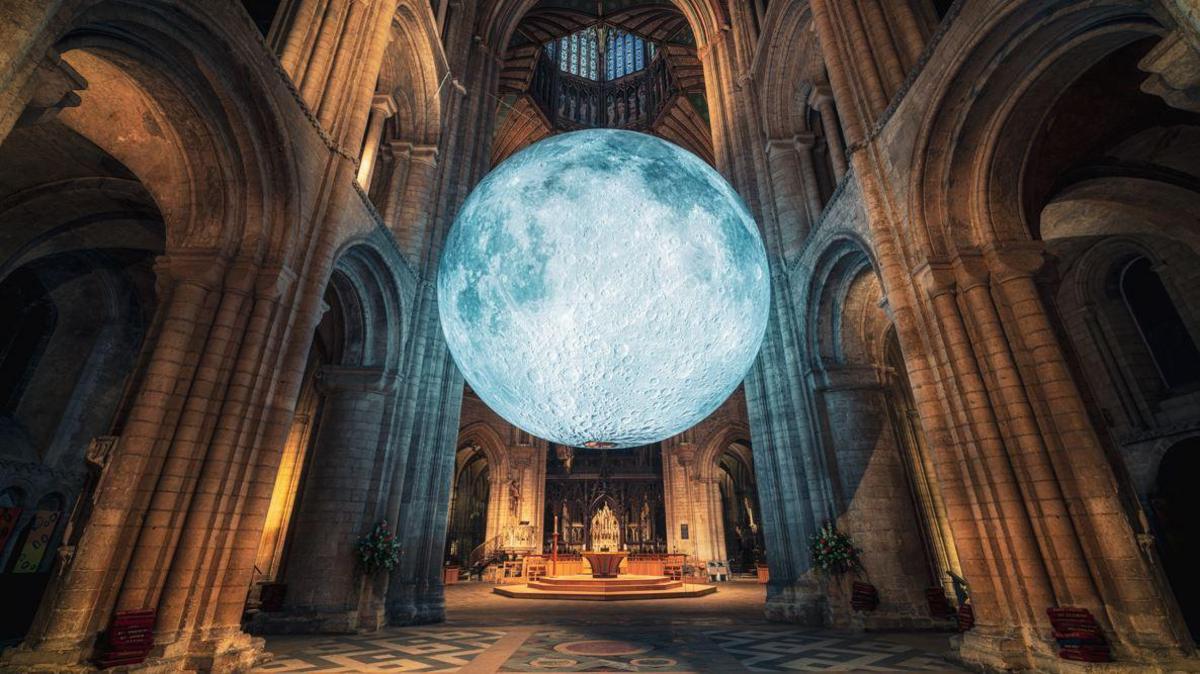 The large grey sculpture of the Moon hangs in front of the altar in a cathedral in between brown sandstone pillars and arches.