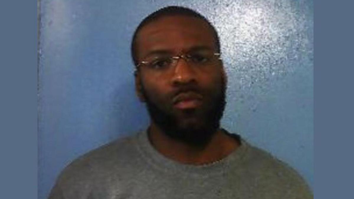 A police custody image of a man in a grey prison tracksuit. He is wearing glasses, a grey prison tracksuit and standing in front of a blue wall.  