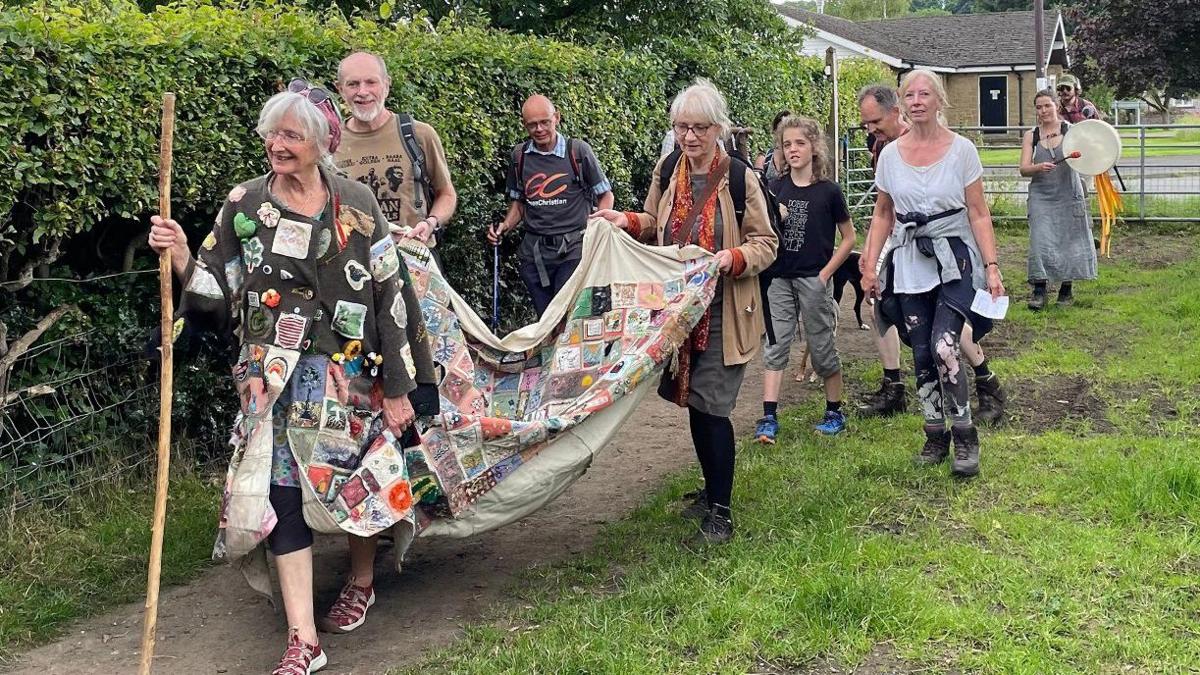 A parade of people in a rural area with the lead walker wearing a large patchwork coat