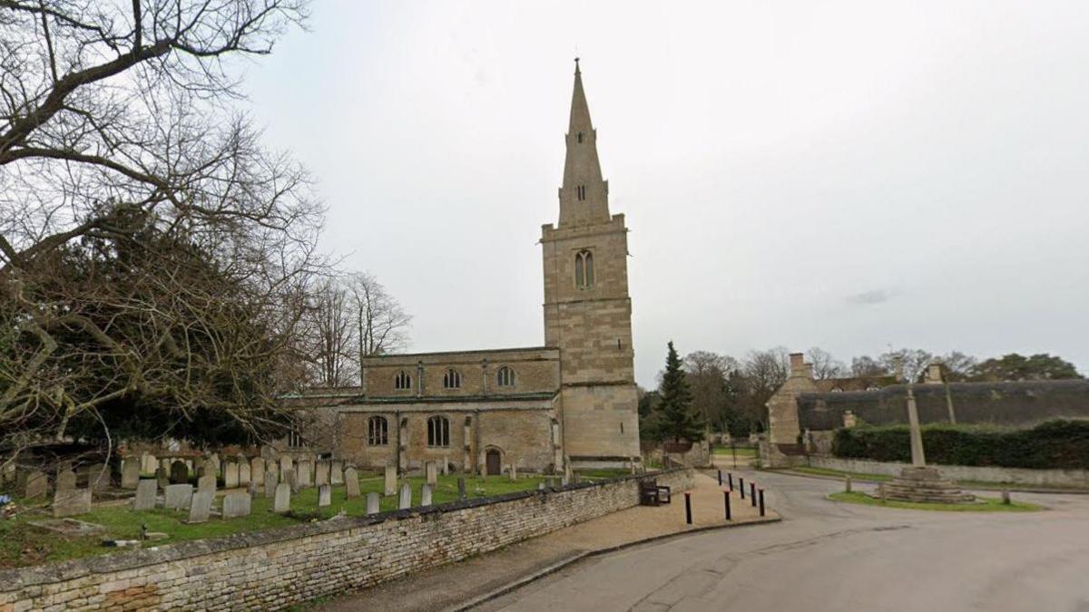 A google streetview shot of a church with a tower and a graveyard in front of it. A road runs alongside the church.