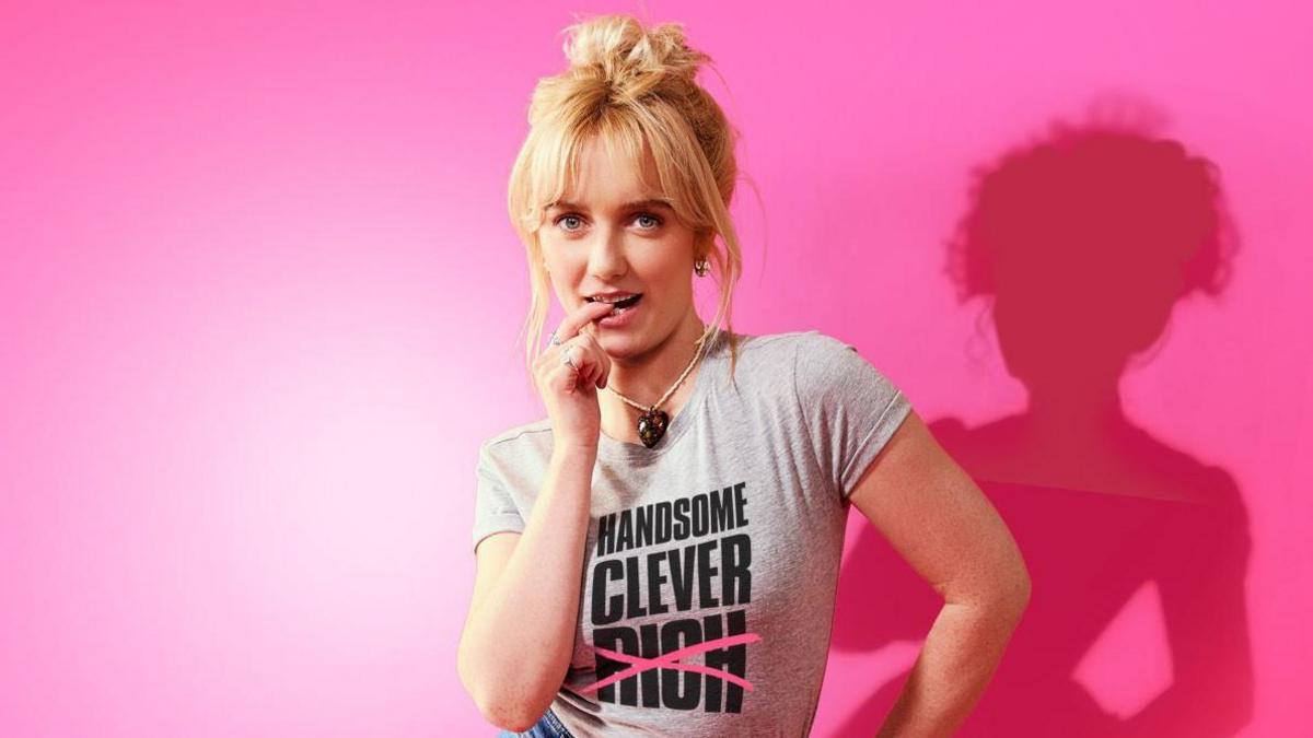 Amelia Kenworthy as Emma wearing a grey T-shirt with her blonde hair piled up in a bun.
