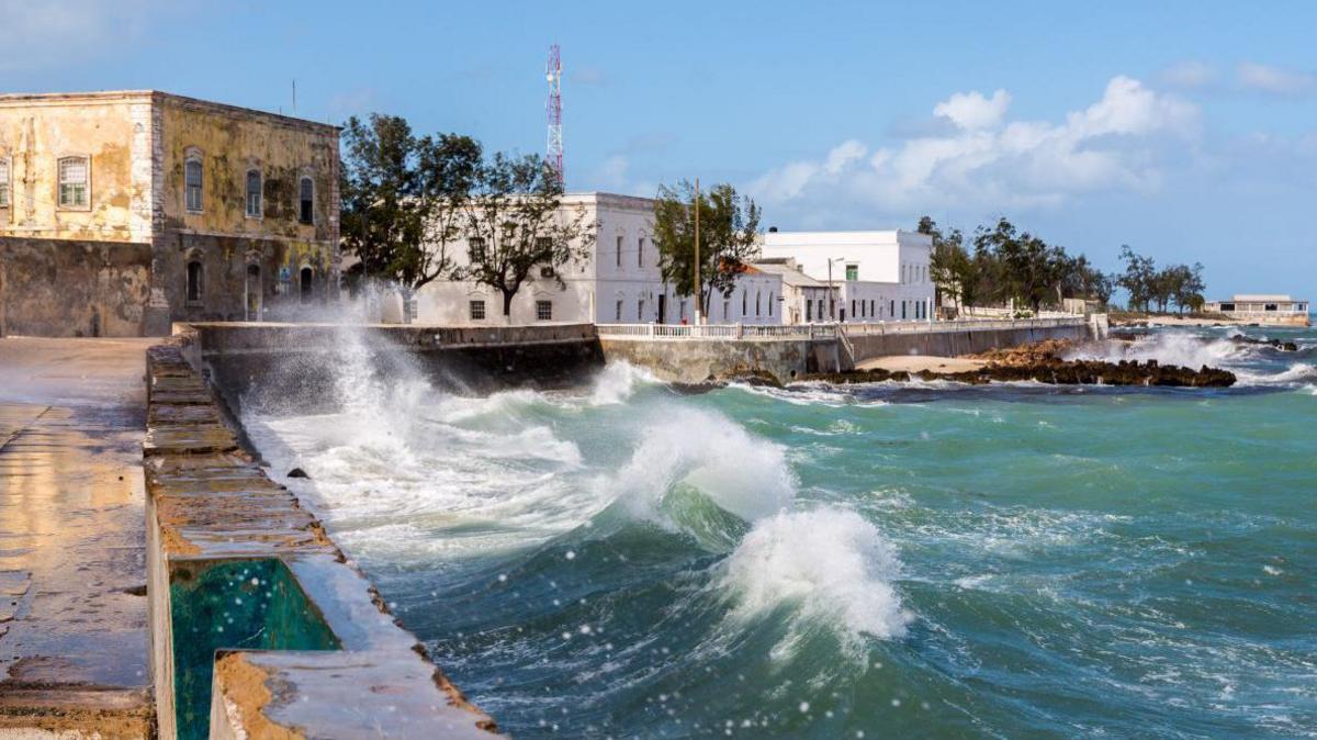 The blue sea crashing against the wall along the Swahili Coastline. There is a brown building on the left hand side and directly in front of the camera there are several white coloured buildings.