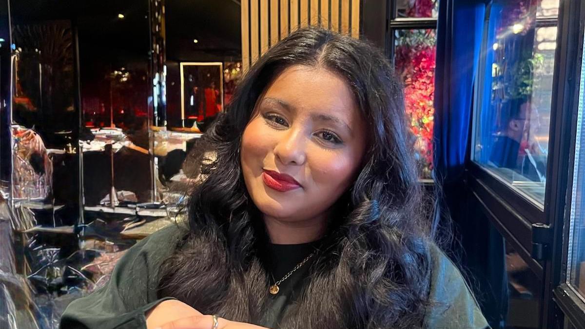 A photo of Hannah Begum sitting in a restaurant. She has long black hair and is wearing a black jumper with a golden pendant. 