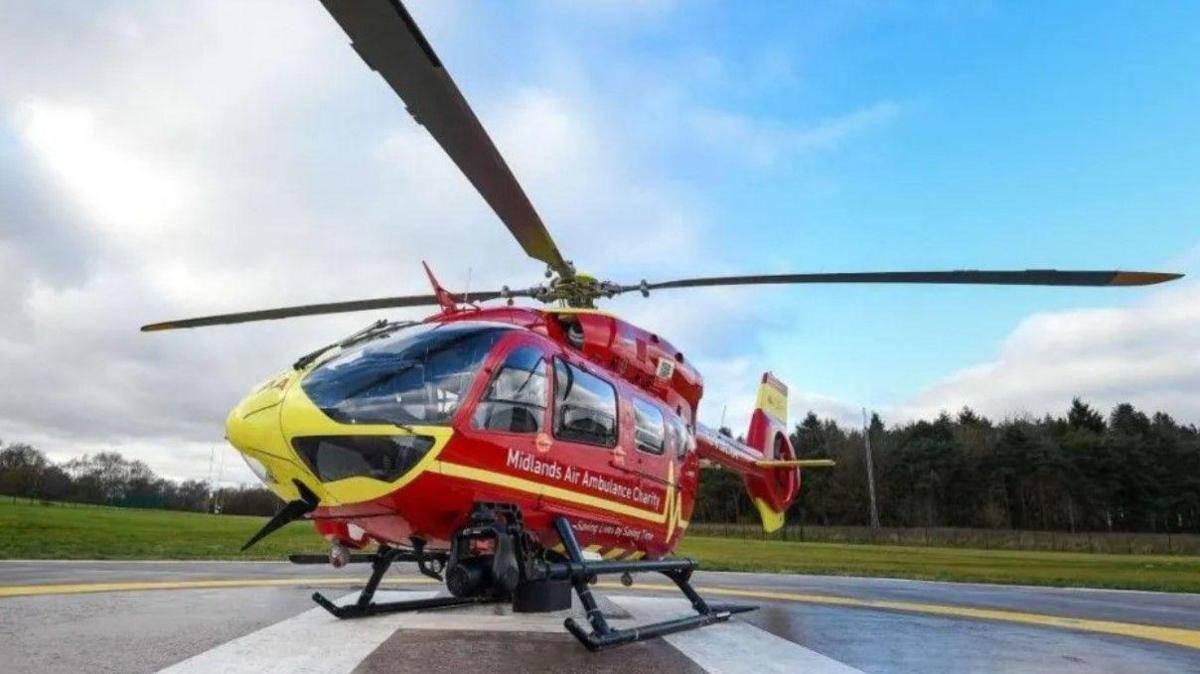 A Midlands Air Ambulance helicopter on a helipad. The helicopter is red and yellow and is pictured on a cloudy day with trees in the background.