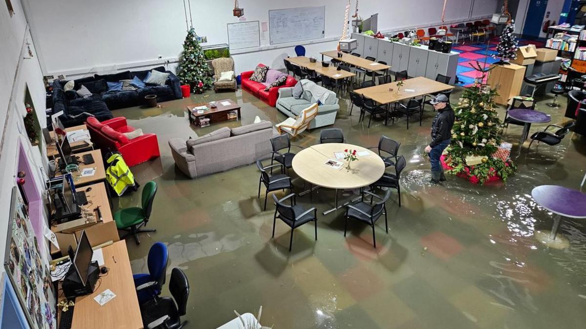 An overhead picture of a large indoor space containing sofas, tables and chairs being flooded at ankle depth. A Christmas tree is also visible, and the water is a muddy brown colour. Coloured carpet tiles are just visible underneath the water. 