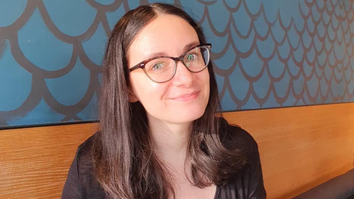 A woman sat down on a bench that is against a wall. She is smiling to camera and is wearing glasses. Behind her is blue wallpaper. 