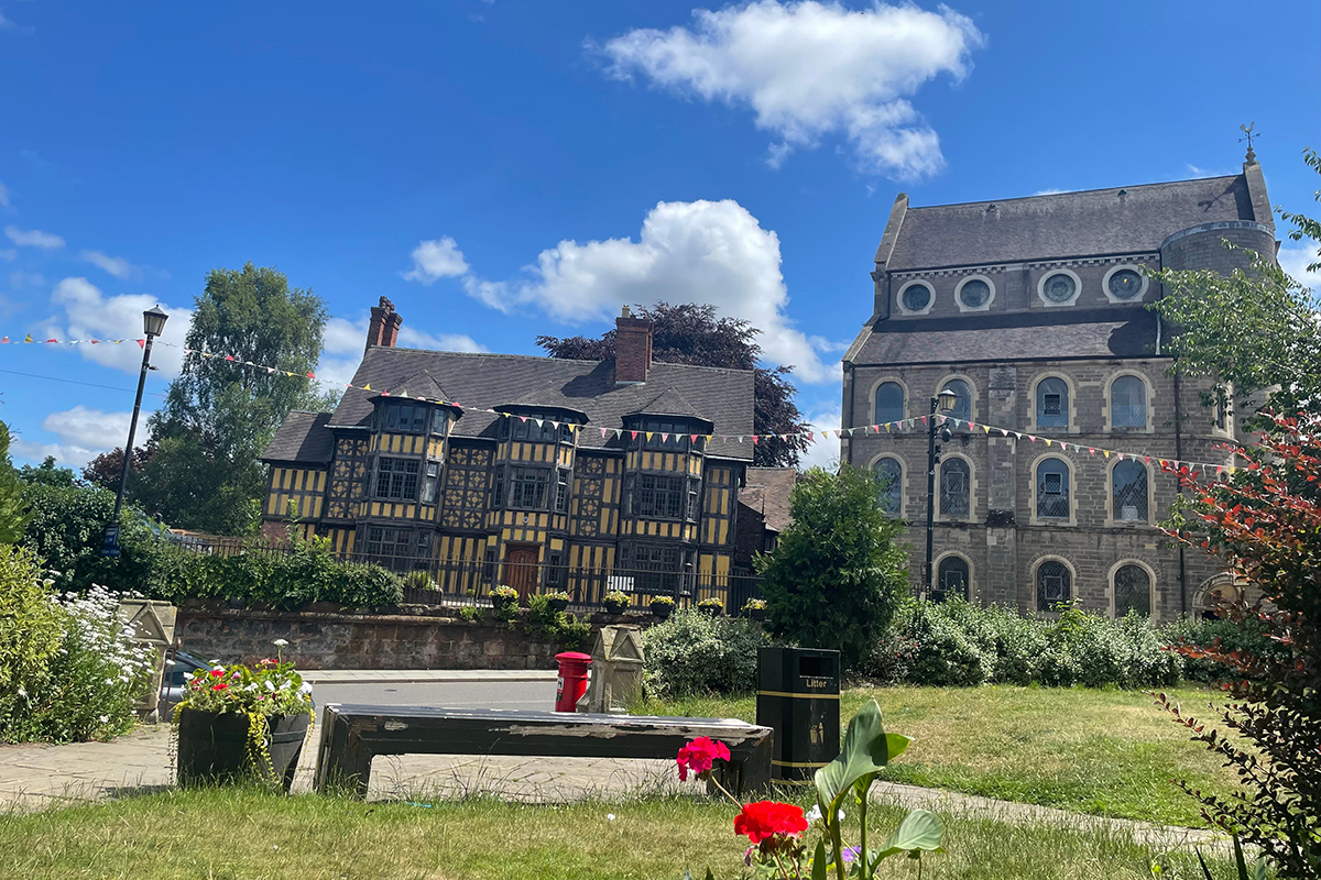 A view of Castle Foregate in Shrewsbury. A bench and pot of flowers is in the foreground on a patch of grass. Across a road is an old timber-framed building in the centre and a large, grey stone-built building on the right