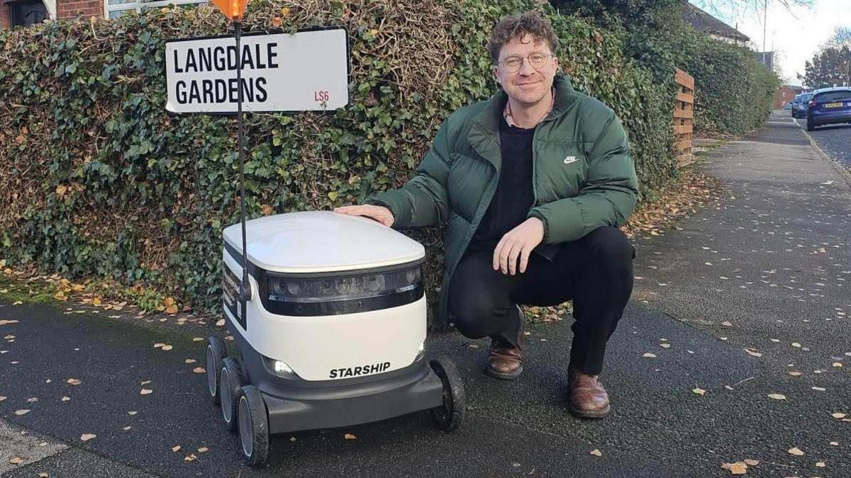 Councillor Jonathan Pryor poses for a photo with a delivery robot. The machine has six wheels and is around 60cm in height. Councillor Pryor has curly strawberry blonde hair and glasses.