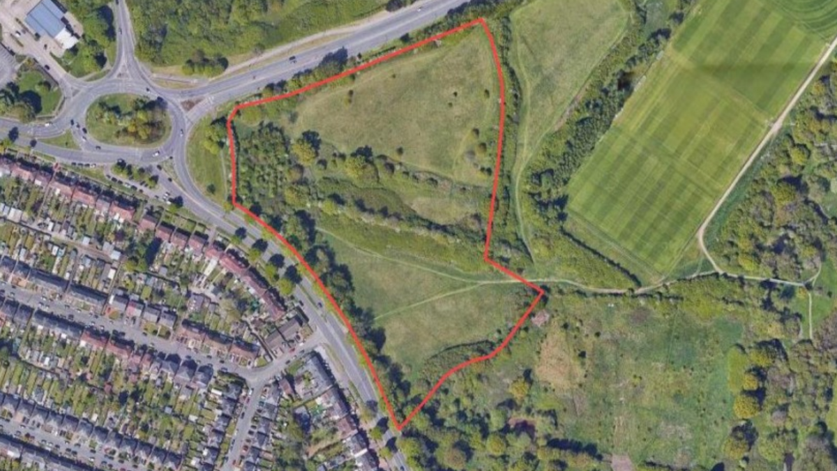 An aerial view of the proposed site, showing green fields to the right of a road and existing housing