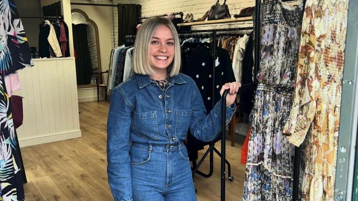 A smiling woman stood holding a clothes railing. She has a blond bob and wearing a denim jumpsuit. The background is a retro style clothing shop. 