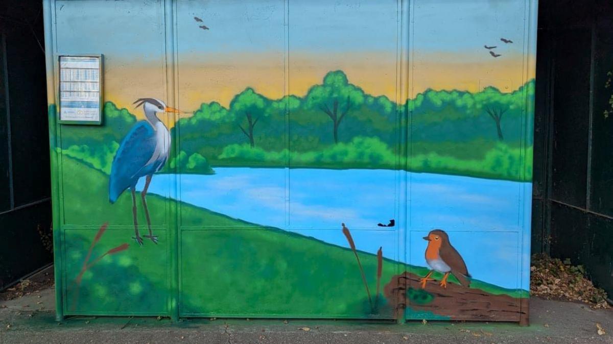A colourful design which features a heron, robin and other birds as well as a blue lake surrounding by green grass and trees. 