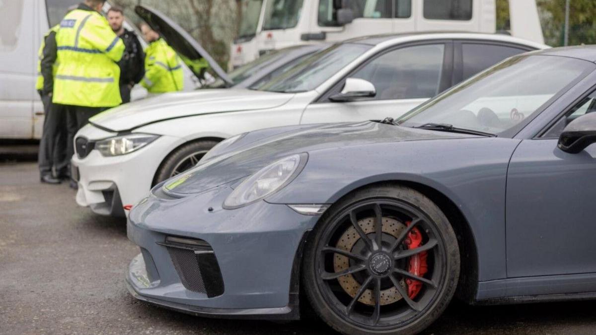 A grey Porsche 911 GT3 in the foreground. A white car, larger vehicles and men are in the background.