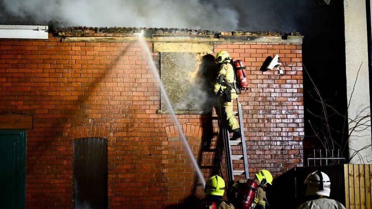 Shot of a firefighter on a ladder tackling the blaze with colleagues below with hoses. There is smoke coming from the red-bricked building and a security camera in view.