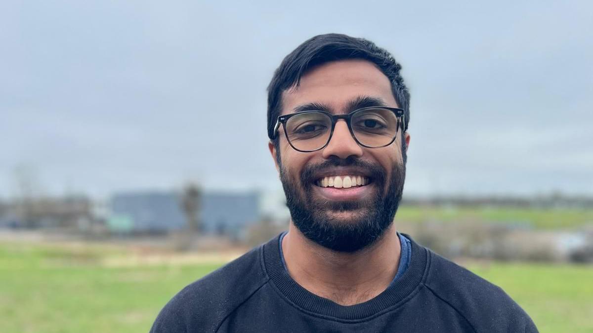 A picture of a man who has glasses on, short black hair and a beard with a moustache. He is wearing a black jumper with a blue t-shirt underneath.