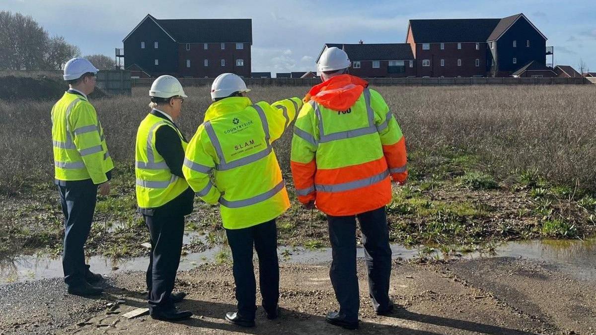 The group of four men in hi-vis jackets, pictured from behind, pointing at a barren piece of land.