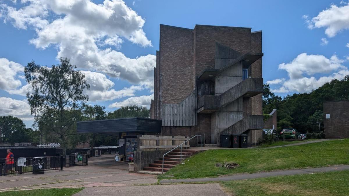 A brown-greyish building is surrounded by green trees and grass. The sky is clear and cloudy. Near the building, there is a small shop.