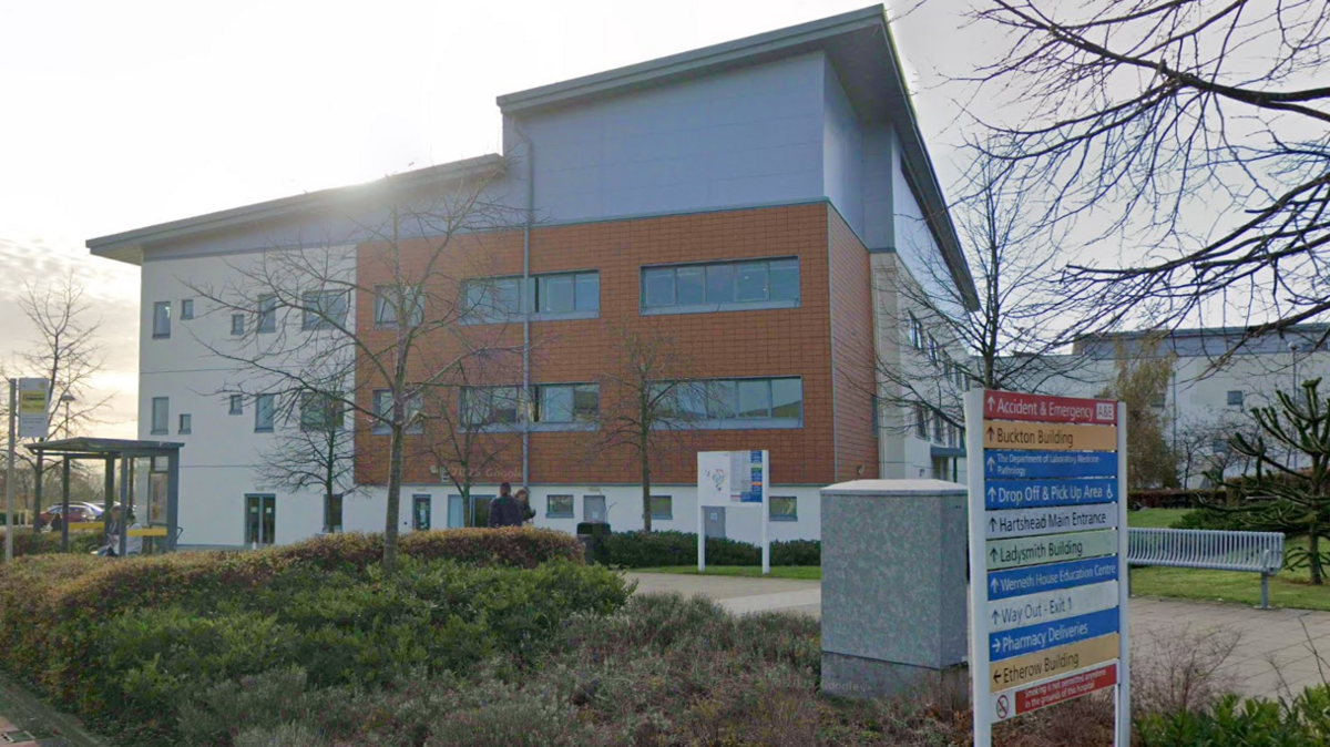 A street view of Tameside Hospital. A portion of the building is made up of red bricks, the rest is a combination of grey and white colours. Outside is a bus stop and greenery.