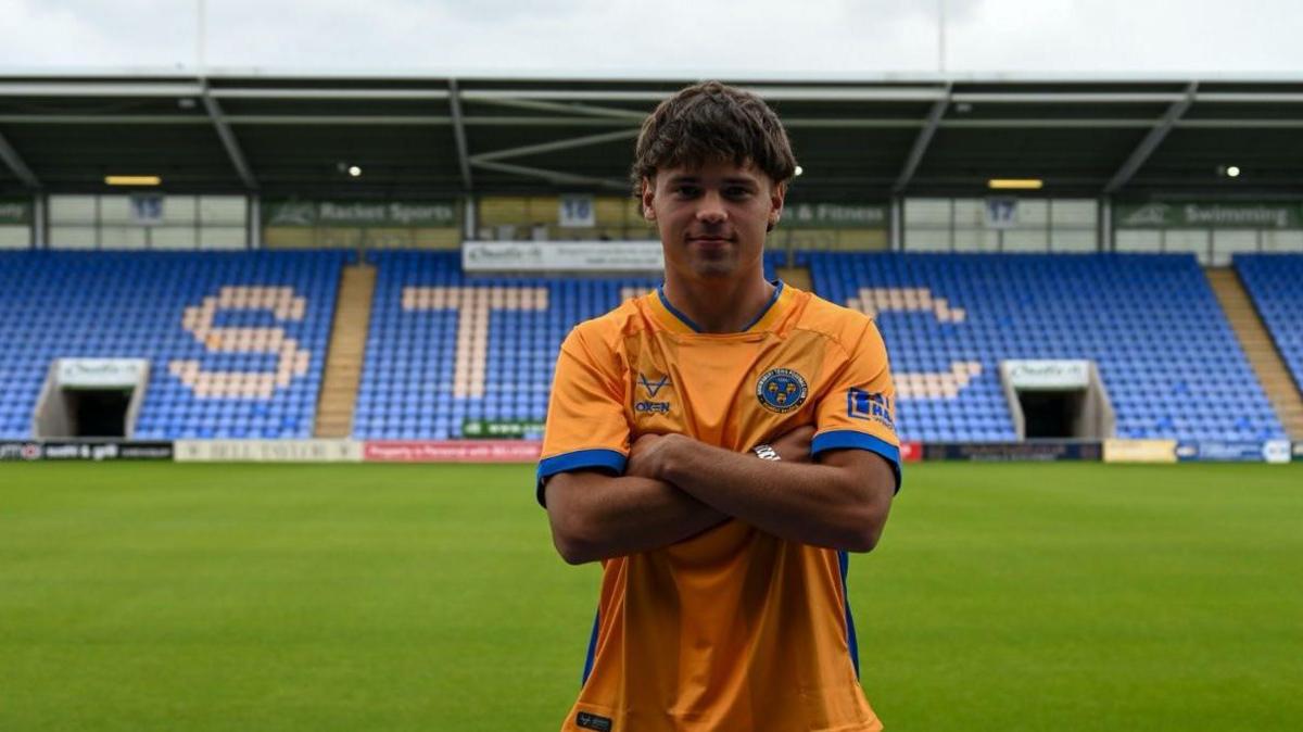 Byron Pendleton folds his arms in a yellow Shrewsbury Town shirt in front of the main stand at Croud Meadow.