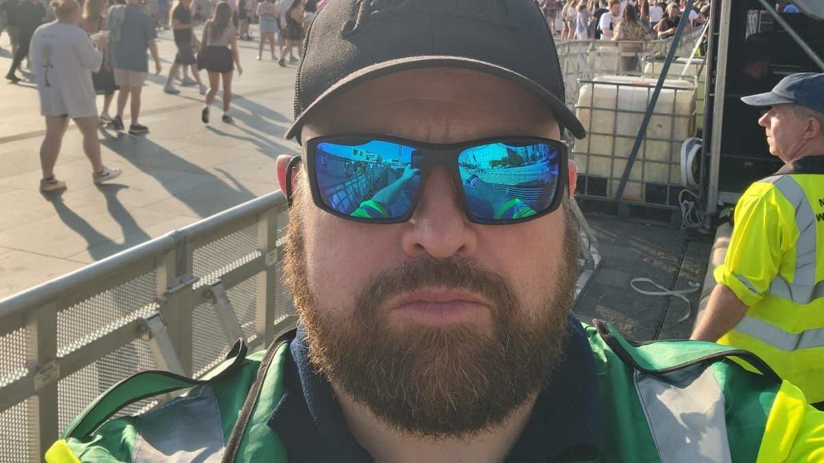 Steve Alcock, who has a ginger beard and is wearing a baseball cap, reflective sunglasses and a green medical uniform, poses for a selfie with the On The Waterfront stage in the background.