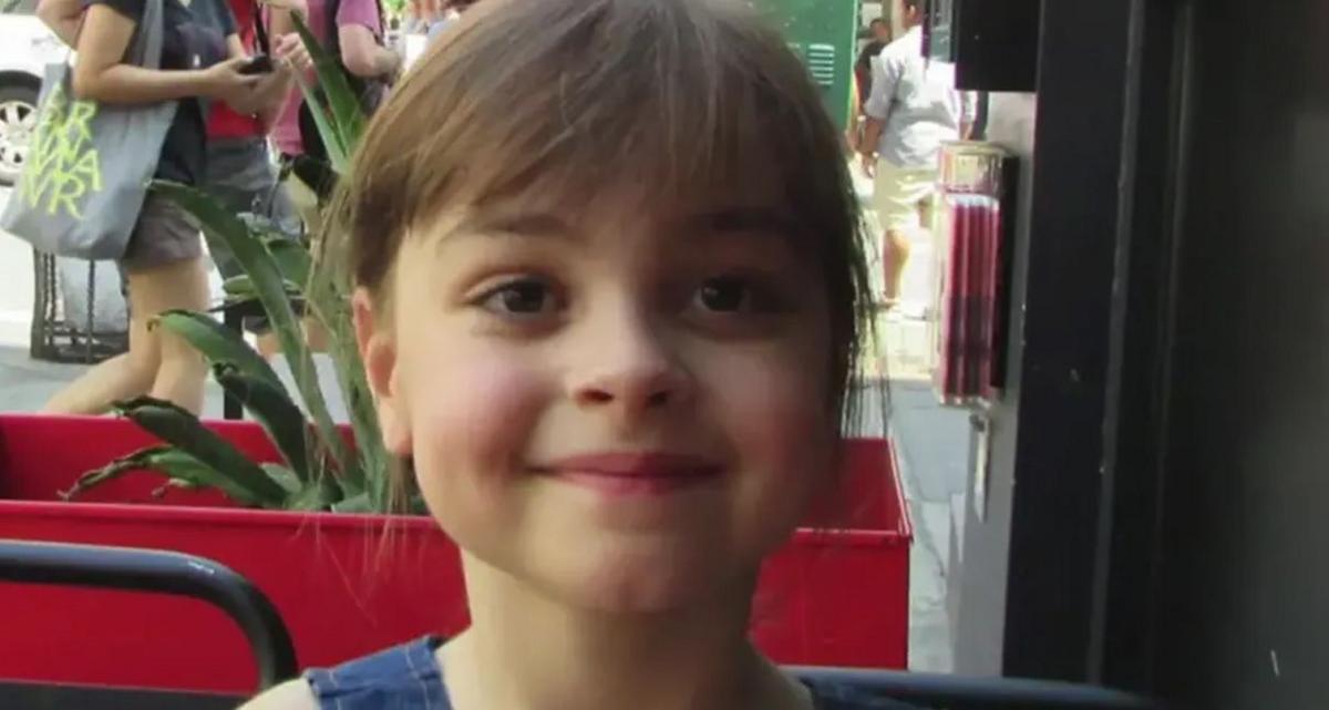 Saffie Roussos sitting outside at a cafe smiling. Her long brown hair with a short fringe is tied up