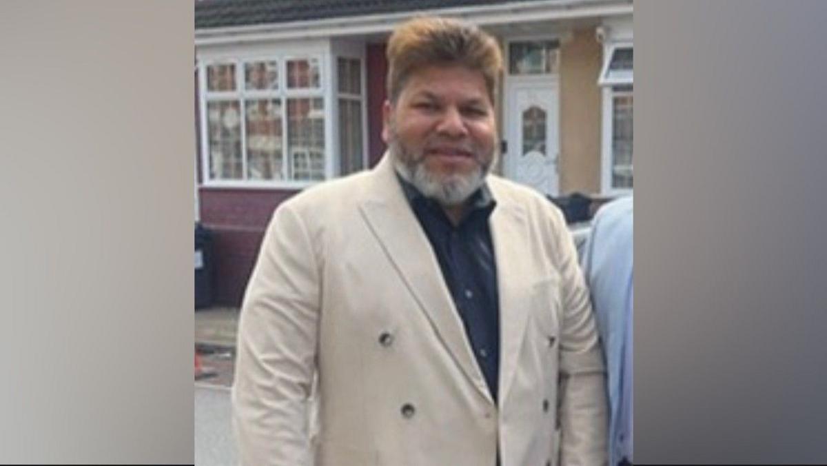 A man with brown hair and wearing a  white jacket over a black shirt, standing on a street in front of a house. He has a grey and black beard.