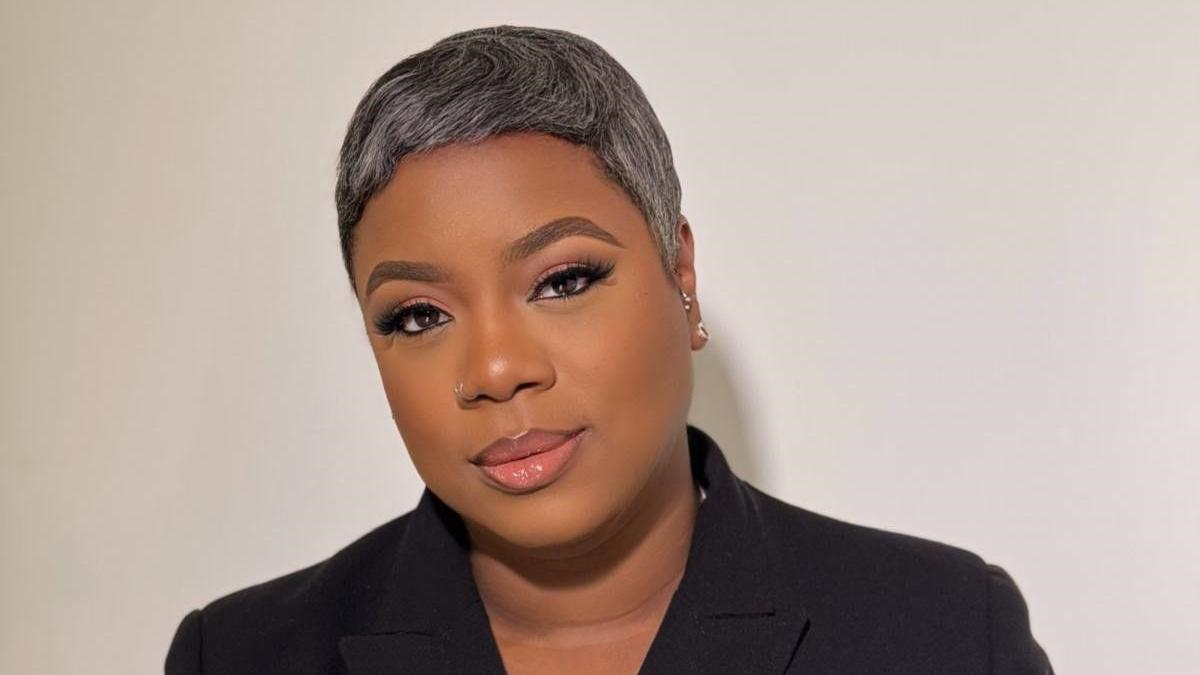 A woman with short dark, grey hair looks into the camera with a slight smile. She wears a dark top with a black collar.