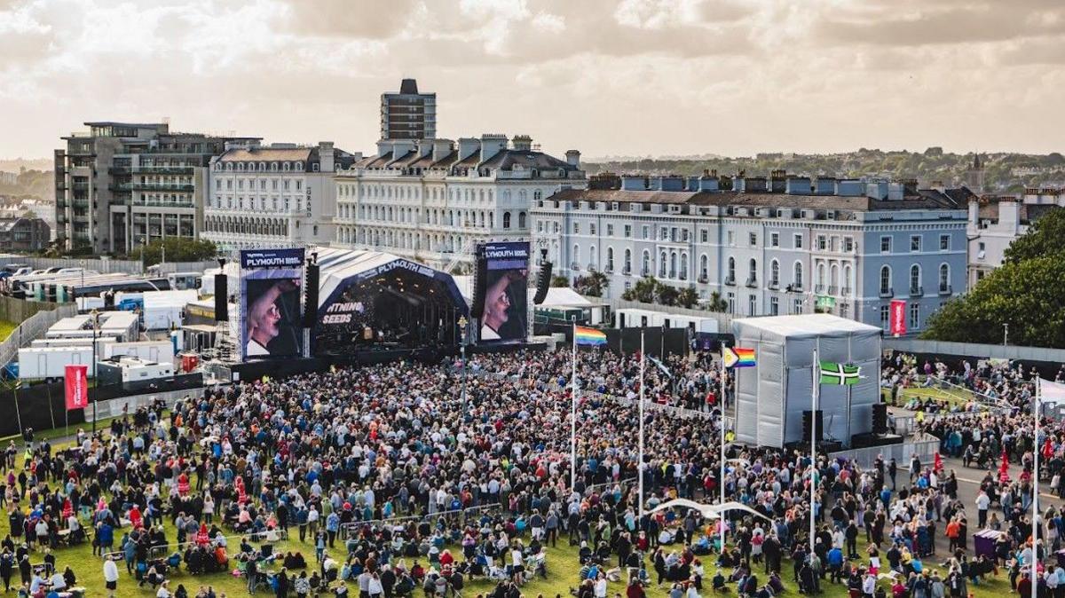 Aerial view of a large crowd at summer sessions in Plymouth. Large crowds have gathered on the Hoe to watch artists on a large stage. Residential properties are located behind the stage. 