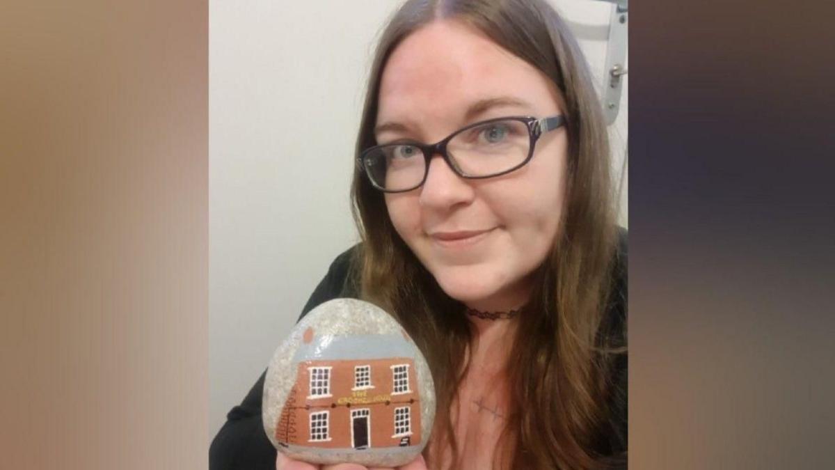 A woman wearing glasses and a black cardigan holding a stone with a painting of the Crooked House pub on it