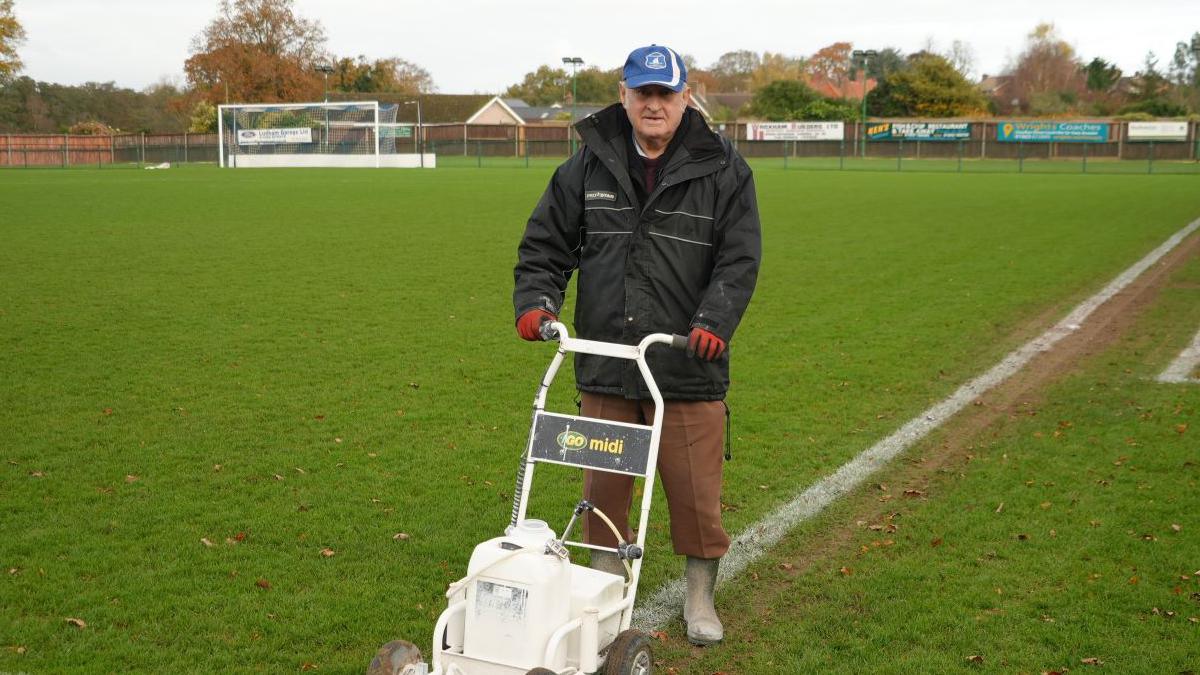 Groundsman Barry Letten wearing wellie boots and a blue cap pushing a line marking machine around the pitch