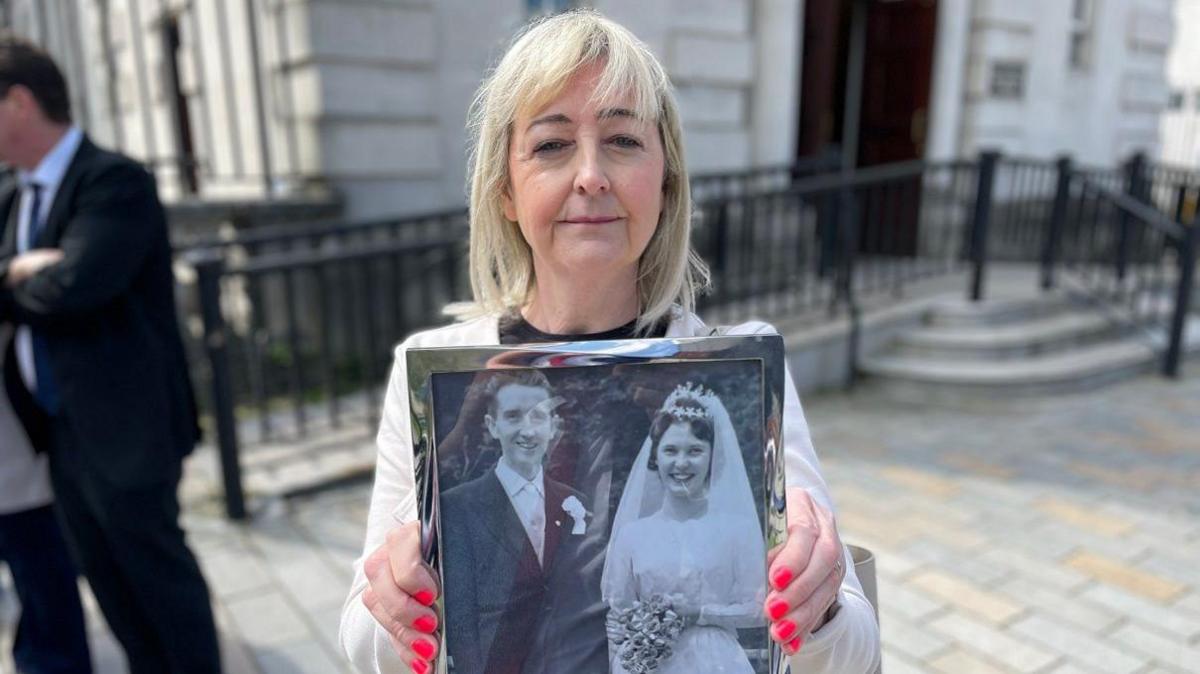 Collette Rooney has light blond hair down to her shoulders and is holding a black and white wedding photograph of her dad and mum while standing outside court