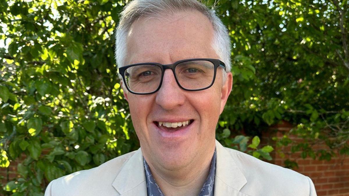 A man with grey hair and wearing a white blazer and black glasses, smiling at the camera. There's a brick wall and a large bush behind him.
