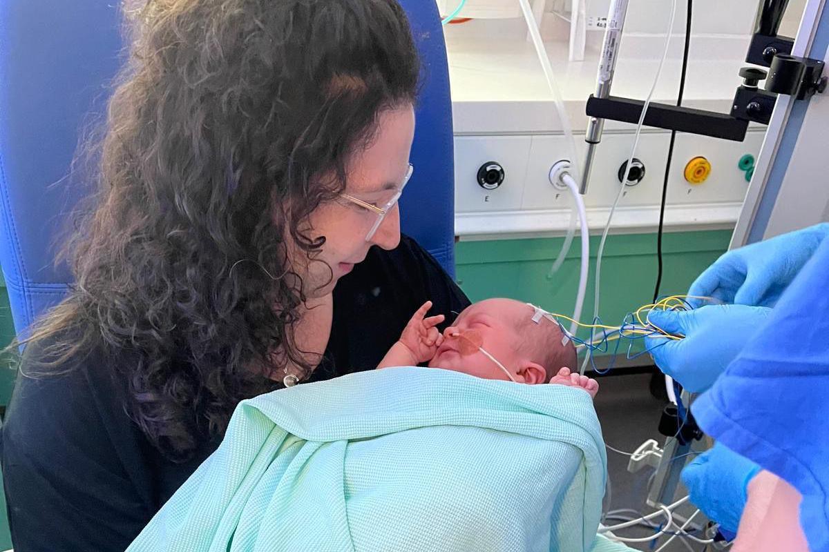 A photo taken in hospital which shows Naomi holding a newborn Roddy. The baby is wrapped in a light green blanket and only his head is visible. It has wires attached to a monitor nearby. Naomi is wearing glasses and looking down at her baby. Also visible in the photo are a pair of blue latex gloves, presumably belonging to a doctor who is caring for Roddy