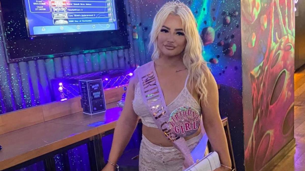 A young woman with long blond hair stands in a bar with her hand leaning on a bar stool. She has a pink sash around her and is wearing a large pink badge which reads 'birthday girl'.