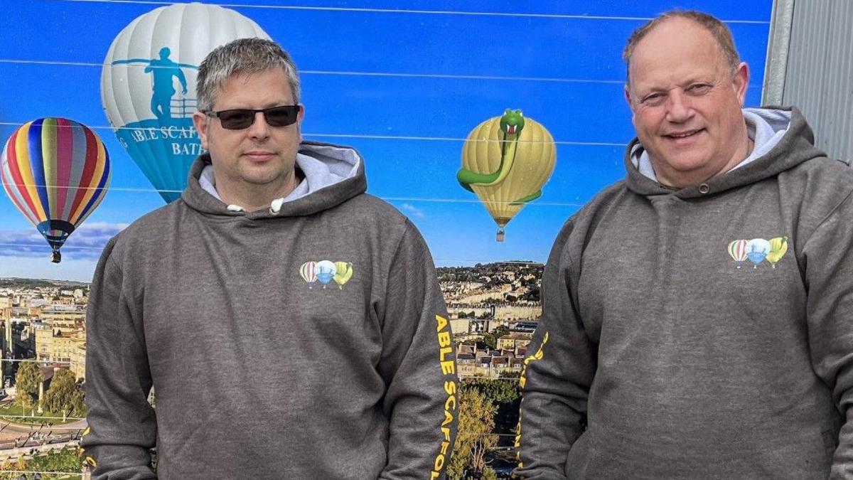 Two men in grey hooded sweatshirts stand in front of an image of hot-air balloons flying over the city of Bath