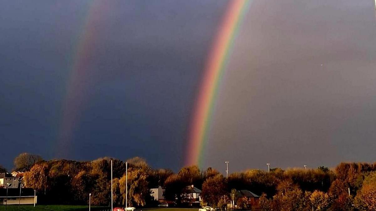 A double rainbow. The rainbow on the right is more brightly coloured. The sky is a dark blue. There are trees which are brown. 