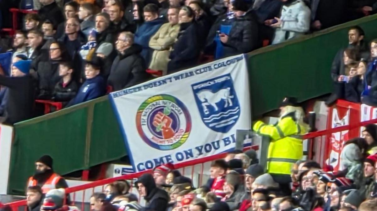 The stands at Portman Road. Fans are sitting in the foreground and standing in the background. The two areas are separated by a green barrier and a red railing. A steward in a yellow hi-vis jacket is standing by the railing. There is a white banner draped over the barrier which says "Hunger doesn't wear club colours".