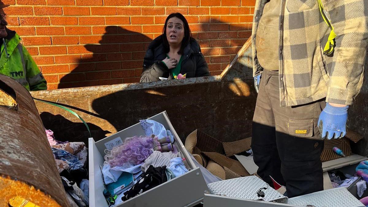 Grace Porter staring at her family's belongings thrown in a skip, including a drawer with toys and notebooks. A man in work clothing stands in the skip.